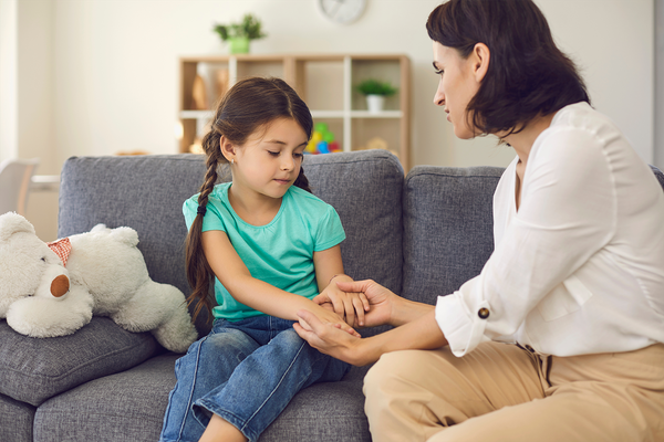 A parent holds a child’s hand while sitting on the couch. The child’s eyes are closed and there is a stuffed animal nearby.