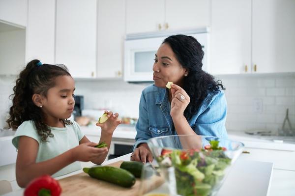 A parent and child sit together while the child tries a bite of a vegetable.