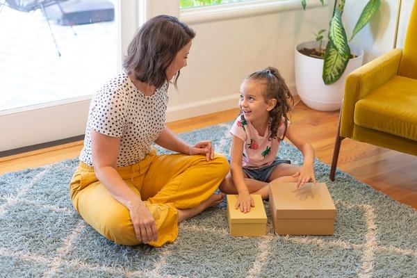 Mother and child sit and sing a song while child is drumming on boxes to make a beat.