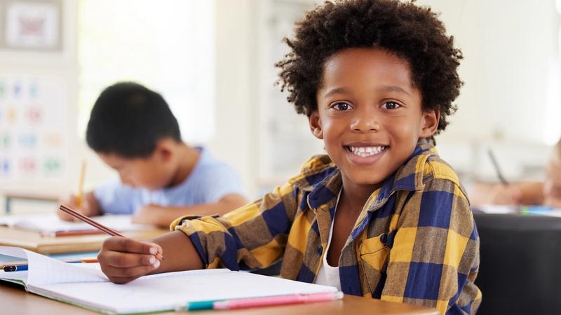 A child smiles while sitting at a school desk holding a pencil and paper.