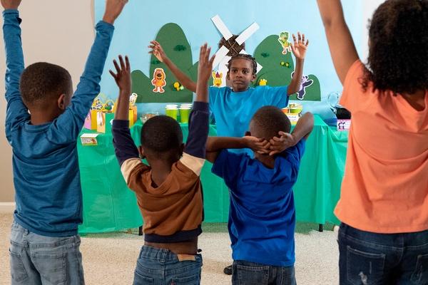 Five children play Donkey Hodie Says. Four children have their arms up in the air.