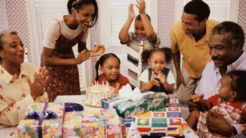 A family is gathered around a table with birthday cake and gifts.