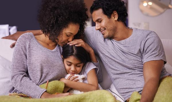 Mother, father, and young daughter snuggling on the couch with a blanket.