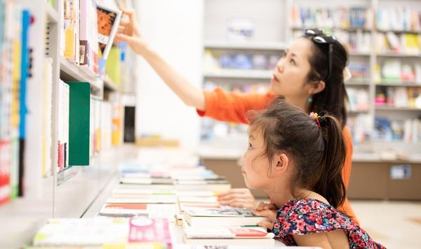 A woman picks up a book from a library shelf while her daughter watches.