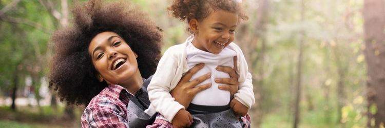 Mother holding daughter while at a park. Both are smiling.