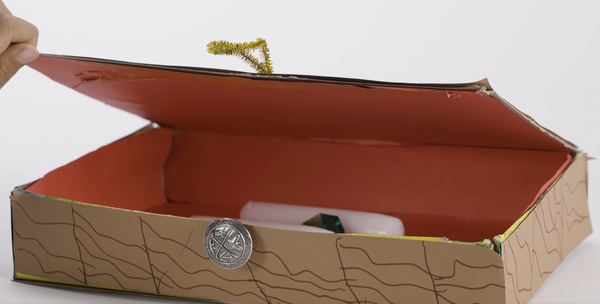 A child opens the lid of a homemade treasure chest made from a cereal box. The words “pirate-themed number hunt” appear on the screen.