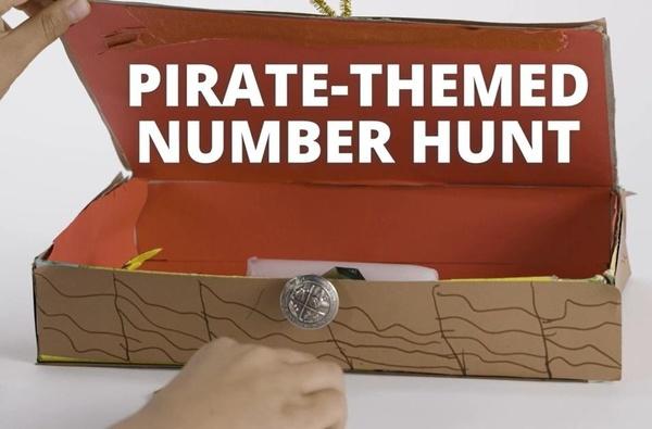 A child opens the lid of a homemade treasure chest made from a cereal box. The words “pirate-themed number hunt” appear on the screen.