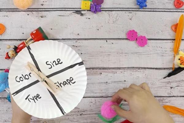 A person holds a paper plate spinner with the words “color, size, type, shape” and a clothespin that can spin between the words. Various objects such as toys, buttons, and pom poms are on a table in the background.