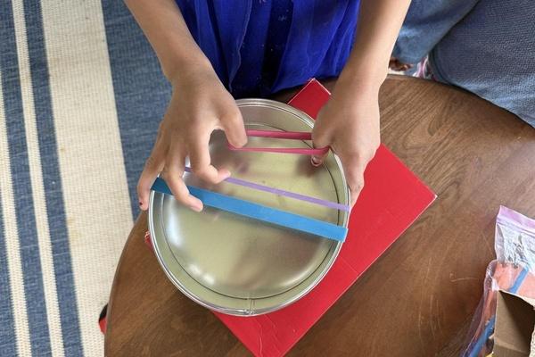 A child places rubber bands on a tin container and then plucks the bands to make sounds.