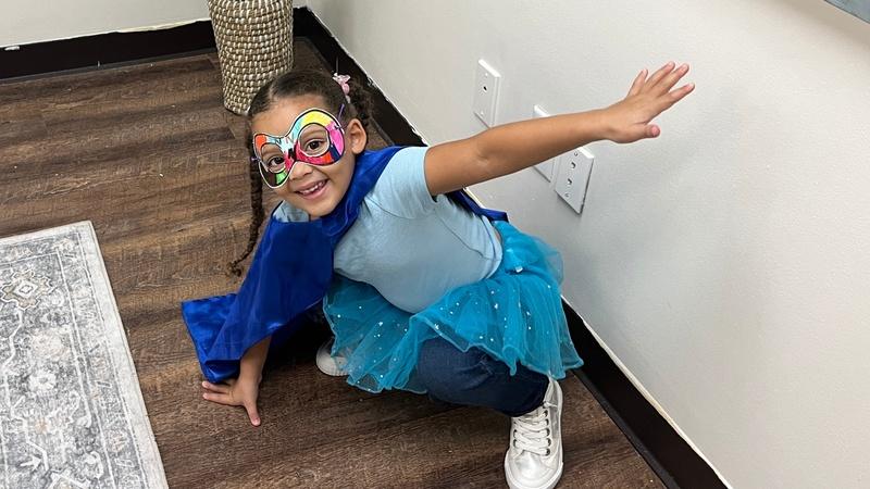 A child is ready to go on a Super Reader adventure wearing a superhero costume including a cape and her Super Reader mask.