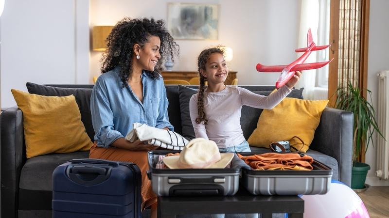 A mother and daughter sitting on a couch getting ready for a trip. There is an open suitcase with clothes on the table in front of them. The girl is flying a toy airplane.