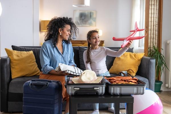 Una madre y su hija están sentadas en un sofá preparándose para un viaje. Hay una maleta abierta con ropa sobre la mesa frente a ellas. La niña está haciendo volar un avión de juguete.
