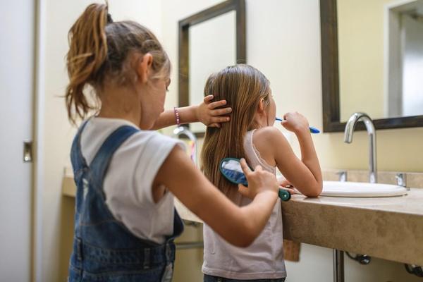 Una niña se lava los dientes mientras otra niña le cepilla el cabello.