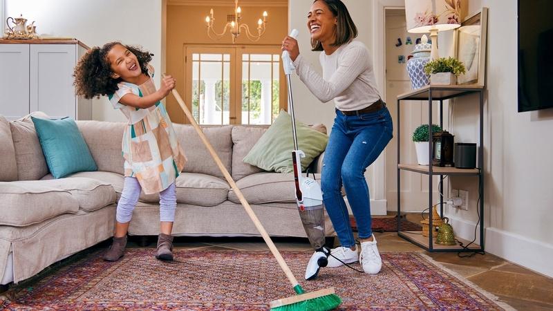 Mother and daughter using vacuum and broom as microphones to sing while cleaning living room.