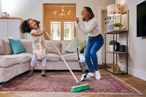 Mother and daughter using vacuum and broom as microphones to sing while cleaning living room.