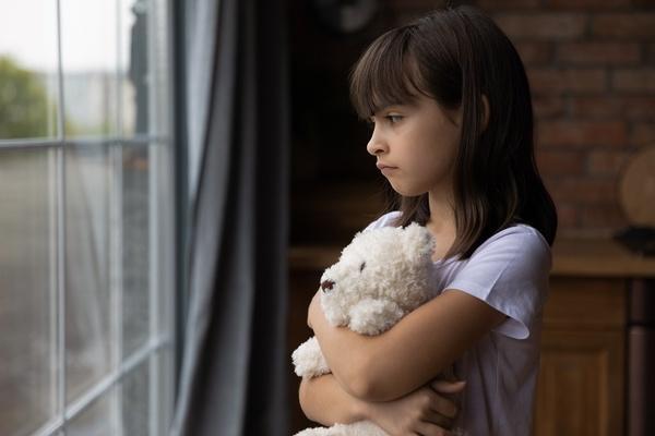 A child who looks sad is holding a stuffed animal and looking out a window.