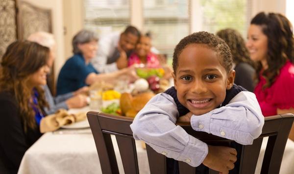 Child sitting at a large dinner table with lots of family members. Child is looking directly at the camera while the family is blurred in the background.