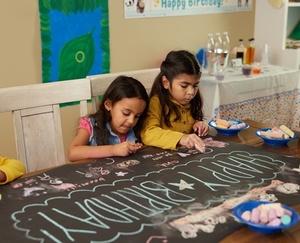 Two children draw with chalk on a birthday banner.