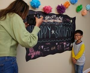 Mom taking a photo of a party guest standing next to the party banner.