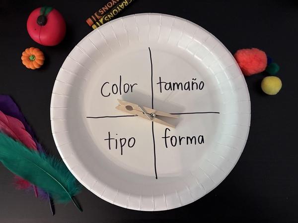 A person holds a paper plate spinner with the words “color, size, type, shape” and a clothespin that can spin between the words. Various objects such as toys, buttons, and pom poms are on a table in the background.