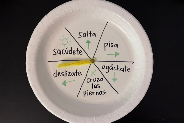 A homemade spinner sits on a table with sections labeled with instructions like jump, step, duck, criss-cross, slide, and shake.