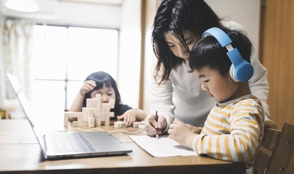 Una madre ayuda a su hijo que está usando una laptop, mientras en el fondo su hija pequeña juega con bloques.